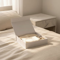 Open silk sleep set gift box with pillowcase, eye mask, and scrunchie on a bed with a marble nightstand